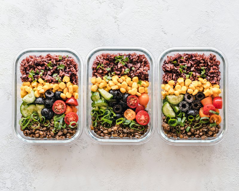 Three glass containers packed with red rice, sweet corn, cucumber, black olives, cherry tomatoes and lentils, portioned for the week.